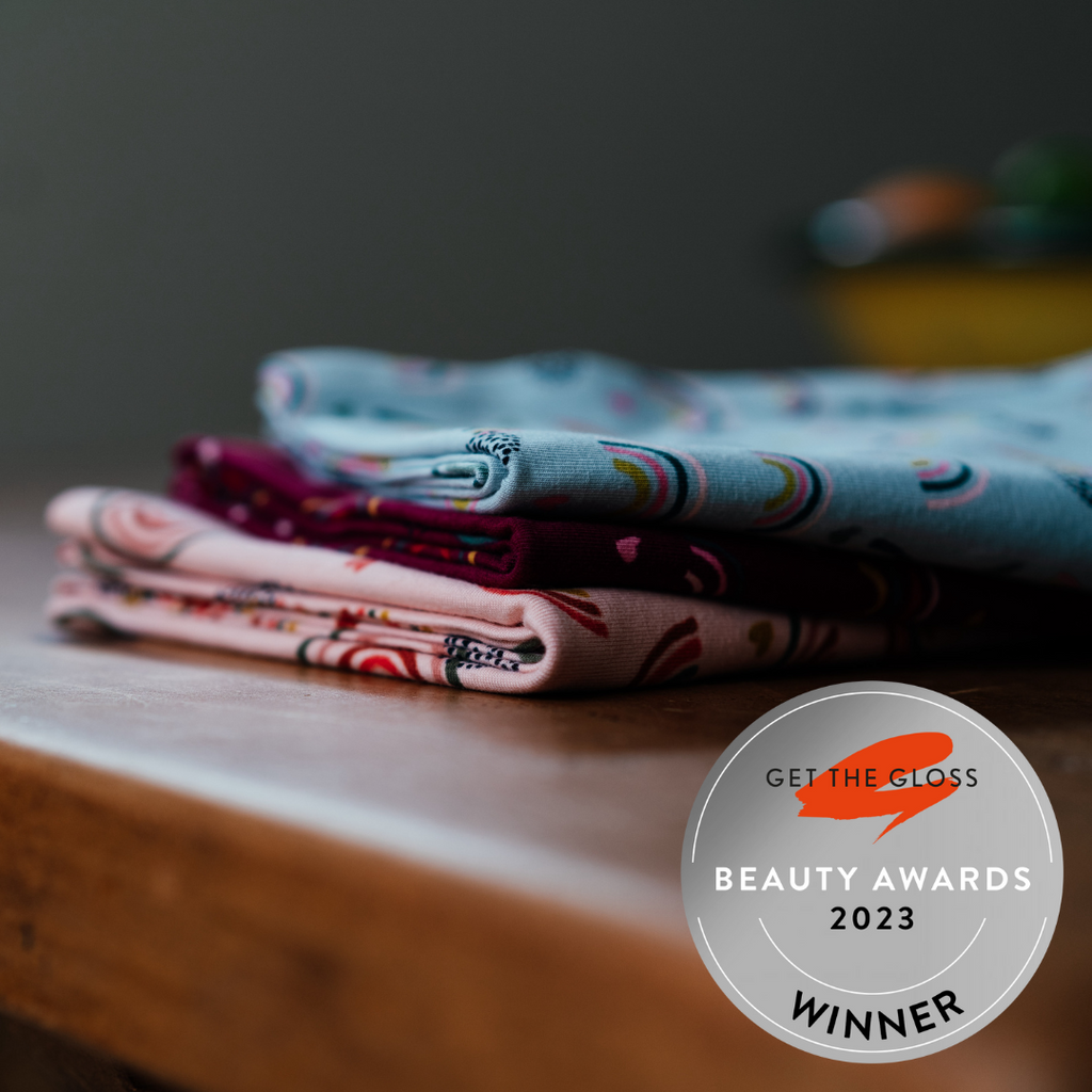 A stack of folded patterned fabrics, featuring The Rainbows organic jersey cotton towel by Good Wash Day, suitable for sensitive skin, on a wooden surface with a "Get The Gloss Beauty Awards 2023 Winner" badge in the bottom right corner.