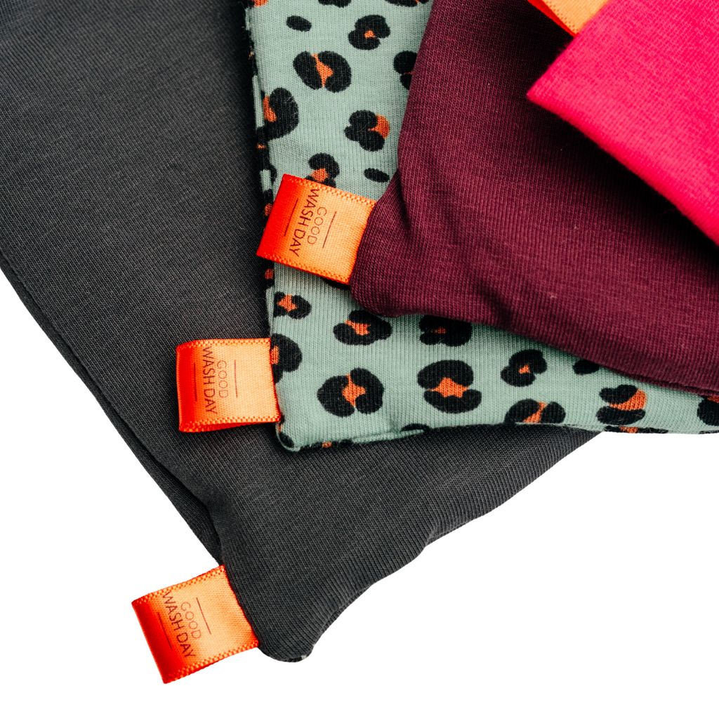 Four folded fabrics with orange tags are stacked; from top to bottom, the colors are pink, burgundy, leopard print, and dark grey. These reusable face cloths from Good Wash Day's "Reusable Face Cloths - Pack of 5 + Bag - Large" collection are made from organic jersey cotton and are perfect for sensitive skin.