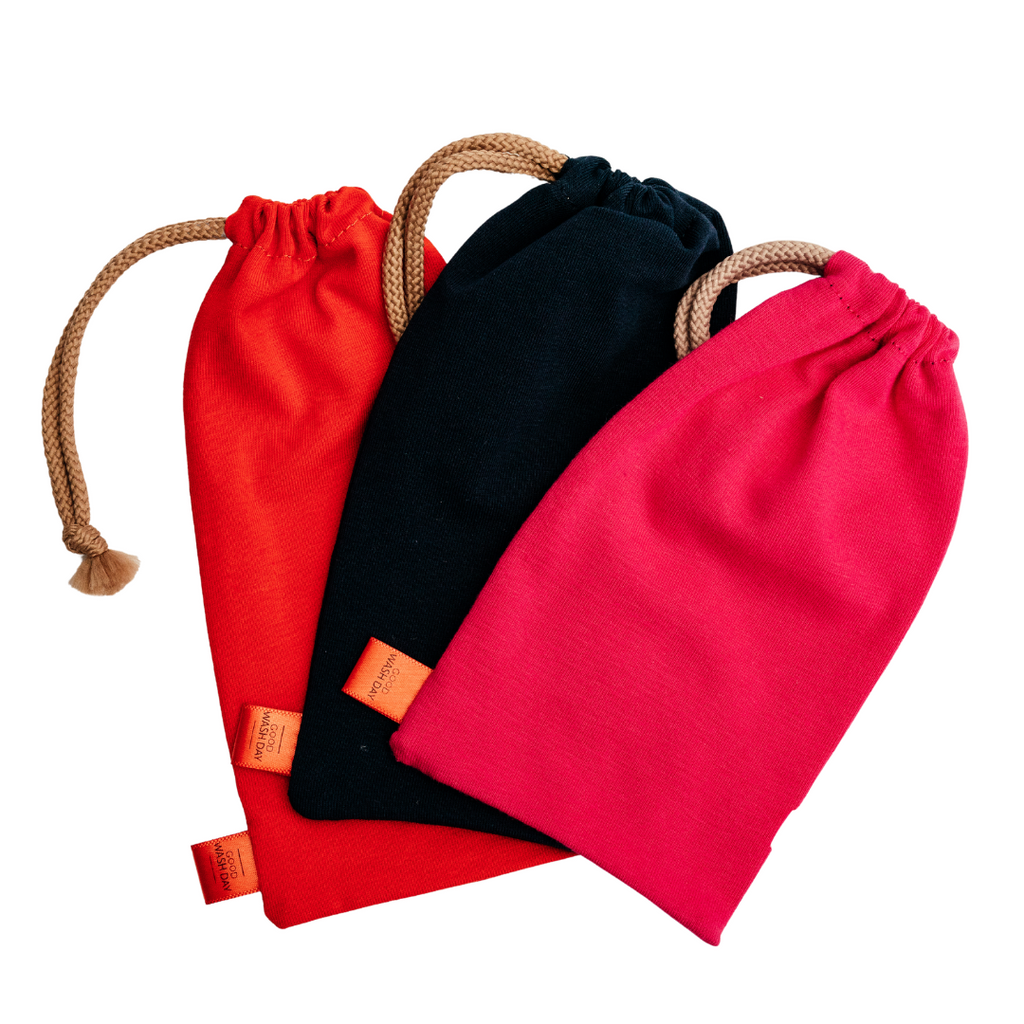 Three Large Drawstring Bags from Good Wash Day, available in red, black, and pink colors, are placed side by side. Each eco-friendly pouch features a beige rope drawstring and small orange tags. Handmade in the UK from organic jersey fabric, these bags also offer a special deal: buy 2 get 1 free.